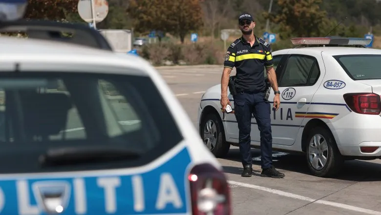 Un polițist stă în picioare lângă o mașină de poliție albă pe un drum asfaltat.