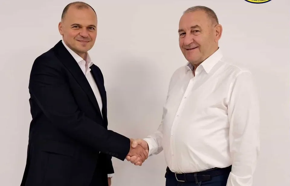 Doi bărbați stau în picioare în fața unui fundal alb și își strâng mâinile într-un handshake.