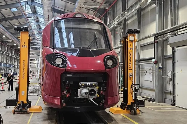 Un tren roșu este poziționat pe șine între două dispozitive de ridicare galbene într-un hangar metalic cu structură gri.