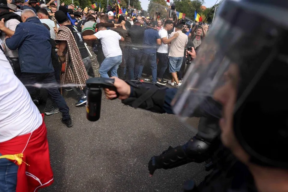Un agent de poliție cu echipament de protecție folosește un dispozitiv de pulverizare spre un grup de persoane într-o zonă stradală aglomerată.