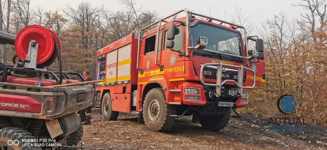 Un camion de pompieri roșu și galben este parcat pe un teren accidentat, lângă un echipament cu un furtun înfășurat pe o bobină roșie.
