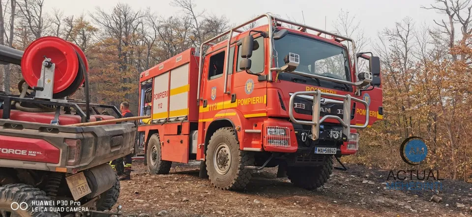 Un camion de pompieri roșu și galben este parcat pe un teren accidentat, lângă un echipament cu un furtun înfășurat pe o bobină roșie.