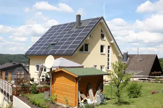 O casă cu un acoperiș înclinat acoperit cu panouri solare fotovoltaice, într-o zonă cu grădină și alte clădiri din vecinătate.