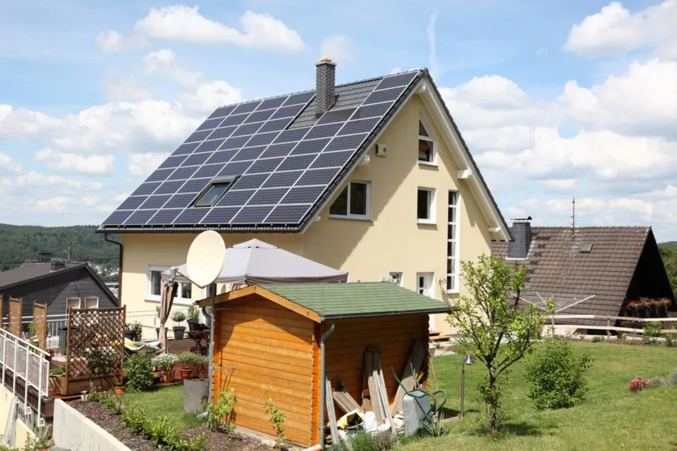 O casă cu un acoperiș înclinat acoperit cu panouri solare fotovoltaice, într-o zonă cu grădină și alte clădiri din vecinătate.
