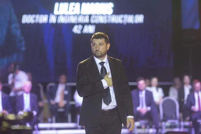 Un bărbat într-un costum închis și cămașă albă stă în picioare pe o scenă în fața unui ecran cu text luminos, în timp ce alți oameni stau așezați în fundal.