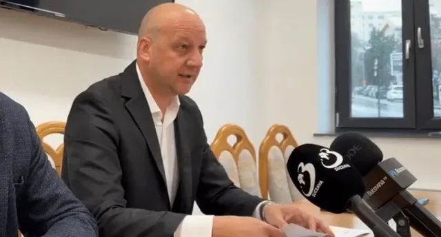 Un bărbat cu o cămașă albă și sacou negru stă la o masă în fața mai multor microfoane cu logo-uri.