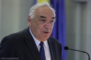 Un bărbat în vârstă cu părul alb, purtând sacou închis la culoare, cămașă albă și cravată albastră, stă în fața unui microfon.