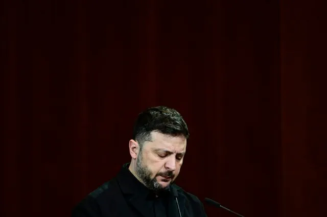 Un bărbat cu barbă și păr scurt, îmbrăcat în haine negre, stă cu privirea în jos în fața unui microfon.