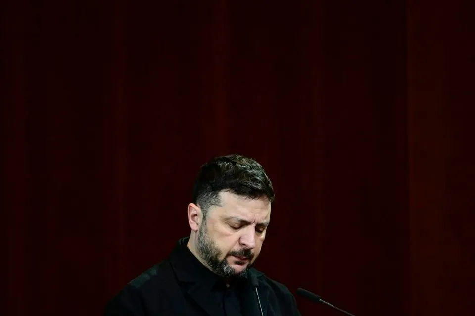 Un bărbat cu barbă și păr scurt, îmbrăcat în haine negre, stă cu privirea în jos în fața unui microfon.