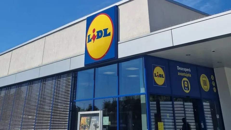 Fațada unui magazin Lidl cu panouri de sticlă albastră și un afiș publicitar pe perete sub cerul senin.