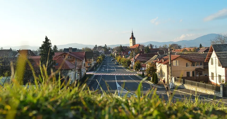 O vedere panoramică asupra unui drum asfaltat care traversează o localitate cu case cu acoperișuri înclinate și un turn de biserică în fundal.