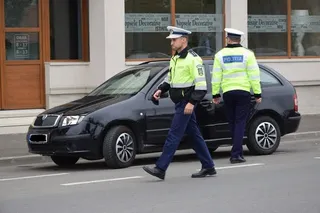 Doi polițiști în uniformă galbenă și albastră merg pe lângă o mașină neagră parcată pe un drum asfaltat lângă o clădire.