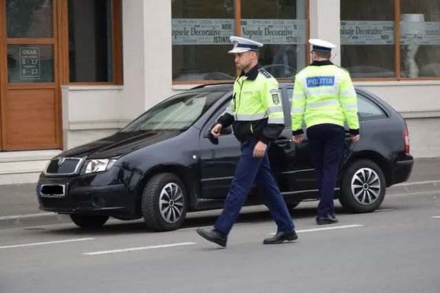 Doi polițiști în uniformă galbenă și albastră merg pe lângă o mașină neagră parcată pe un drum asfaltat lângă o clădire.