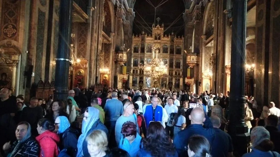 O mulțime de oameni stă în picioare în interiorul unei biserici decorate cu coloane înalte și iconostas ornate.