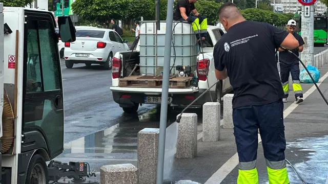 Un bărbat cu tricou negru și pantaloni închiși cu benzi galbene luminoase folosește un furtun de apă pe o stradă umedă lângă un vehicul utilitar.