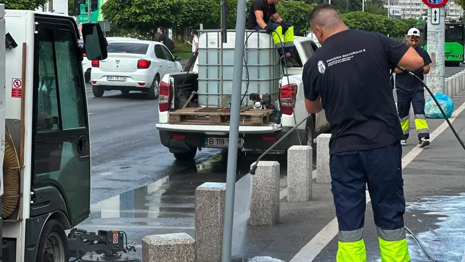 Un bărbat cu tricou negru și pantaloni închiși cu benzi galbene luminoase folosește un furtun de apă pe o stradă umedă lângă un vehicul utilitar.