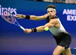 Un jucător de tenis cu bandană albă lovește o minge galbenă cu o racletă albastră și neagră pe un teren de culoare albastră.