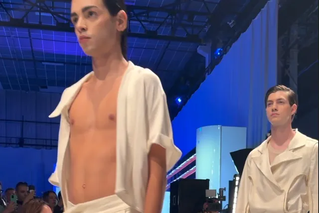 Un bărbat cu pielea deschisă poartă un tricou alb deschis, iar în fundal se observă un alt bărbat cu o cămașă albă sub lumini albastre.