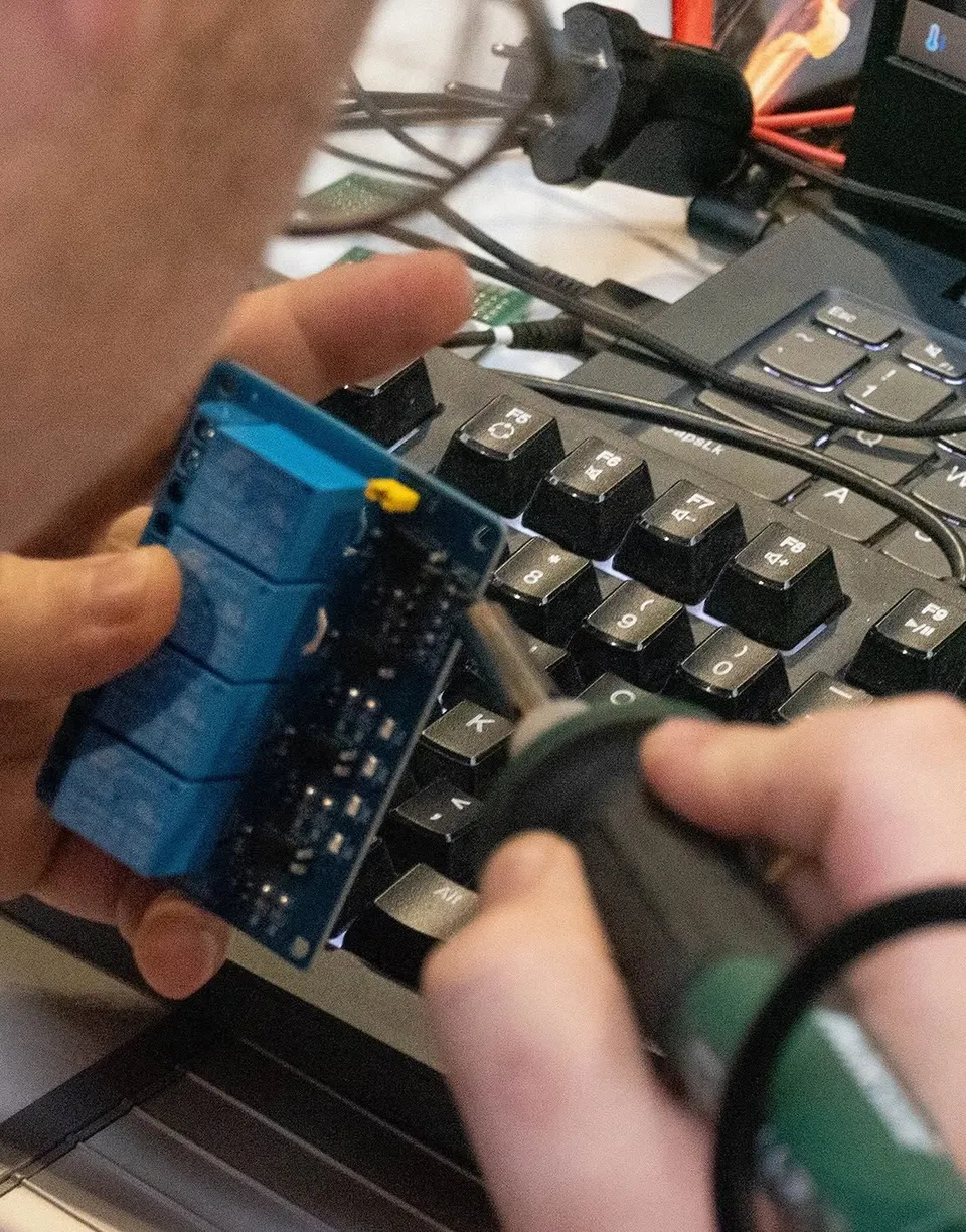 O persoană ține un dispozitiv electronic albastru deasupra unui tastatură de laptop, folosind o unealtă de lipit cu fir de cupru.