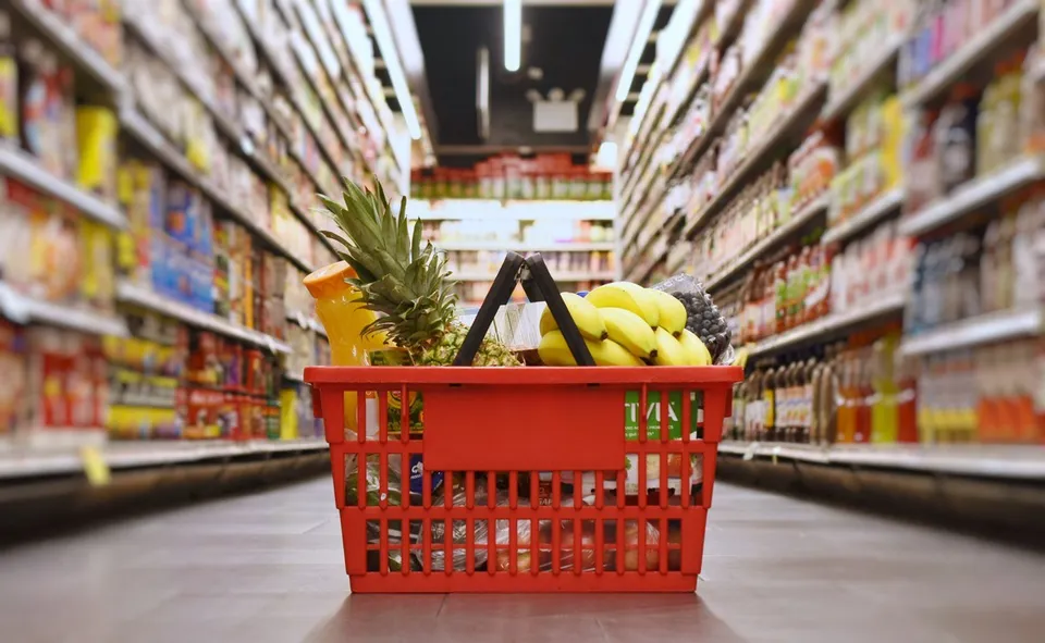 Un coș de cumpărături roșu plin cu fructe, inclusiv banane și ananas, este situat pe un culoar de supermarket între rafturi cu produse alimentare.