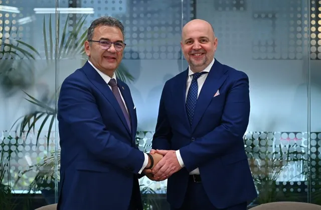 Doi bărbați în costume de culoare închisă stau în picioare și se strâng în mână într-un birou cu plante în fundal.