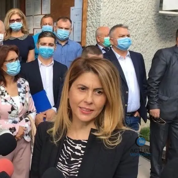 O femeie cu părul blond și bluză cu model alb-negru stă în prim-plan, în fața unui grup de persoane purtând măști de protecție.