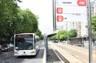 Un autobuz alb cu numărul 323 circulă pe un drum asfaltat lângă o stație de autobuz cu un panou indicativ și un autobuz de tip troleibuz.