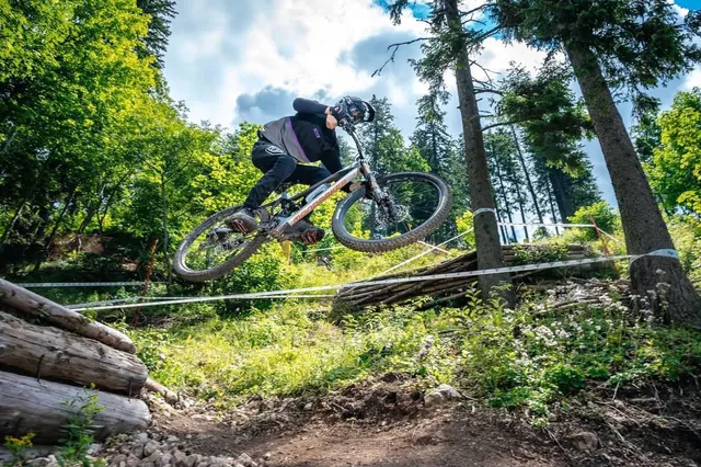 Un biciclist cu cască și echipament sportiv sare cu o bicicletă de munte peste un obstacol într-o zonă împădurită.