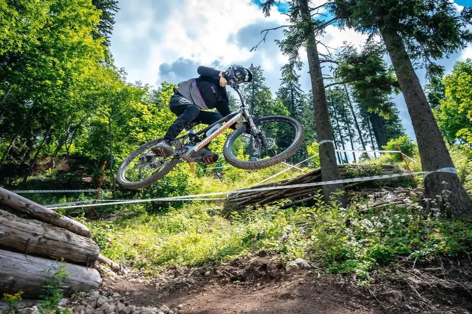 Un biciclist cu cască și echipament sportiv sare cu o bicicletă de munte peste un obstacol într-o zonă împădurită.