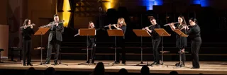 Șapte persoane în haine negre susțin o reprezentație muzicală pe o scenă, folosind flauti și un instrument de suflat sub lumini de spectacol.