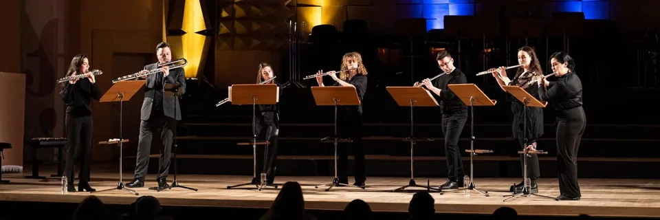 Șapte muzicieni în haine negre susțin un recital pe o scenă iluminată, folosind flautul în fața unor pupitre de partitură.