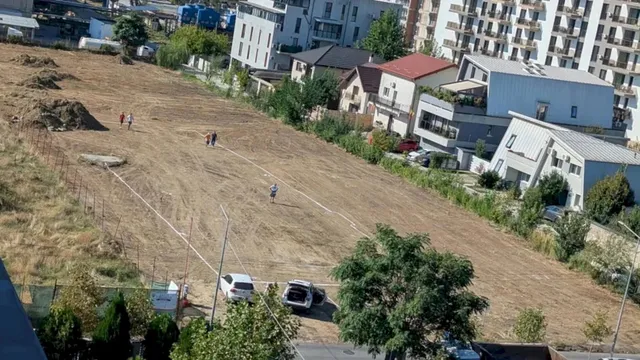 O fetă merge pe un teren gol și uscat, înconjurat de clădiri rezidențiale și vegetație, cu câteva mașini parcate în partea de jos.