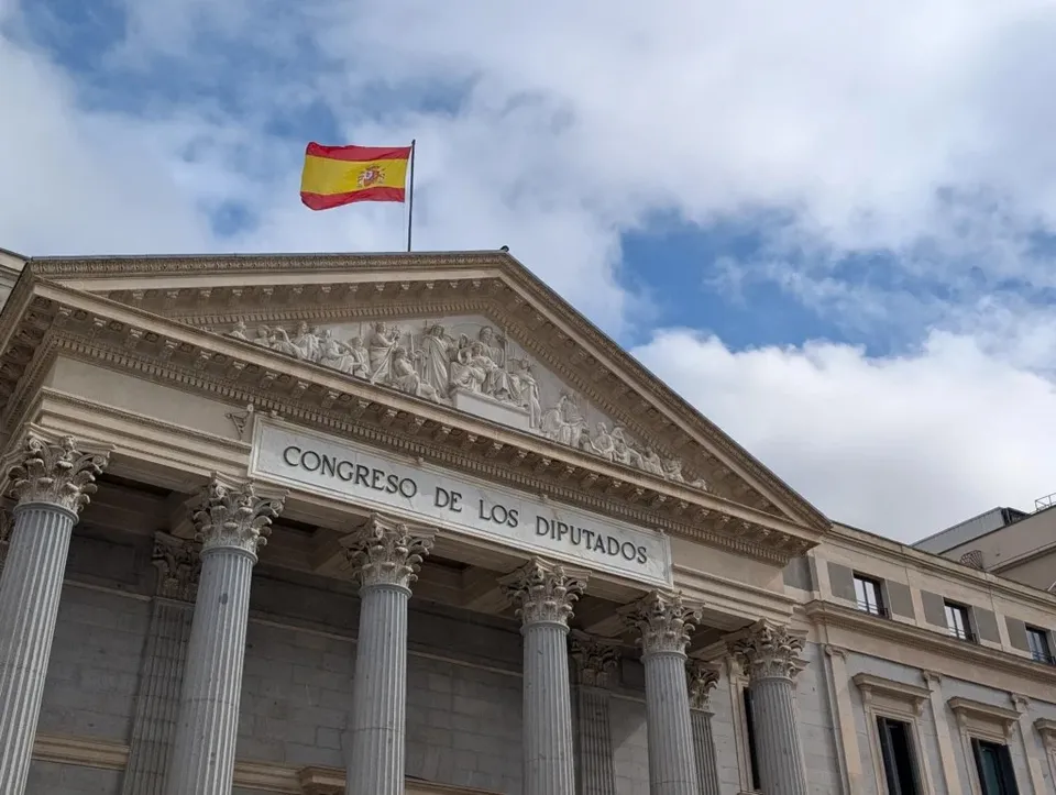 O clădire cu coloane și un fronton sculptat prezintă inscripția "CONGRESO DE LOS DIPUTADOS" sub un steag spaniol agitat pe un cer înnorat.