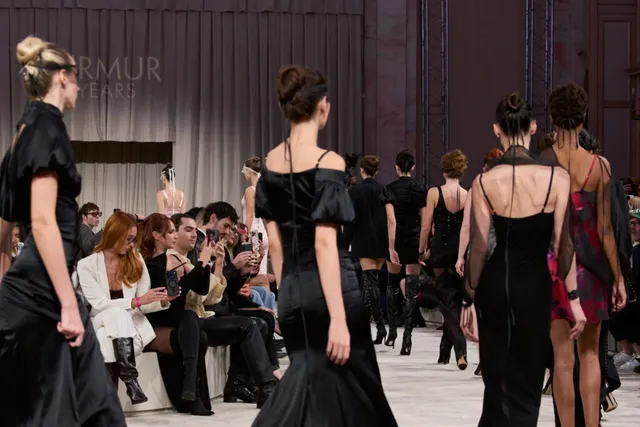 Modele în haine negre merg pe o pistă de catwalk în fața unui public așezat pe scaune.