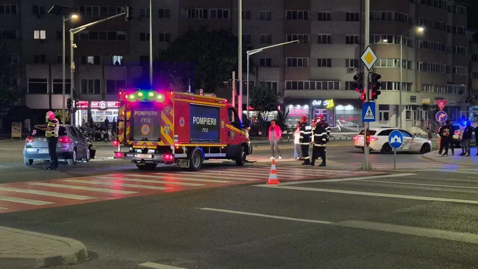 Intersecție nocturnă cu pompieri, polițiști, autoturisme avariate, ambulanță și conuri, în fața unor blocuri iluminate.