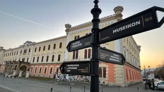 Un stâlp de indicatoare stradale negru este poziționat în prim-plan în fața unei clădiri mari, galben-roz, sub un cer luminos de seară.