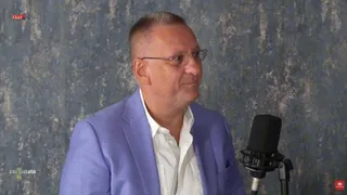 Un bărbat cu ochelari și sacou albastru deschis stă în fața unui microfon într-un cadru cu fundal texturat gri.