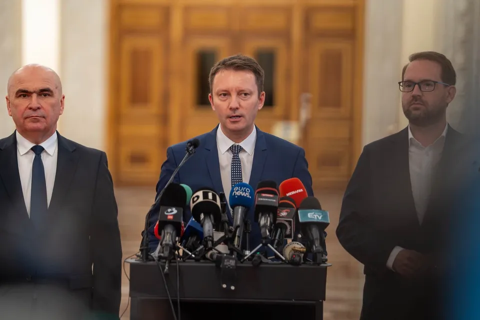 Trei bărbați în costume purtând cravate stau în spatele unui grup de microfoane de presă într-o încăpere cu uși de lemn în fundal.