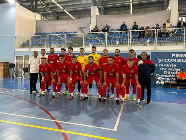 O echipă de sportivi în echipamente roșii și galbenii stă în rând pe un teren de futsal în fața unor tribune cu scaune albastre.
