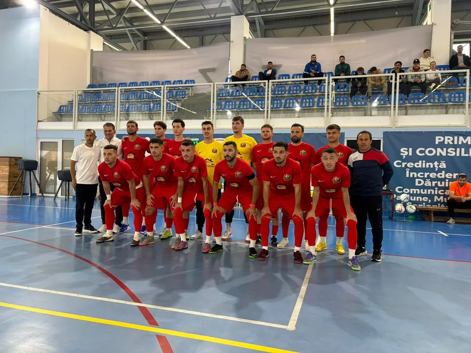 O echipă de sportivi în echipamente roșii și galbenii stă în rând pe un teren de futsal în fața unor tribune cu scaune albastre.
