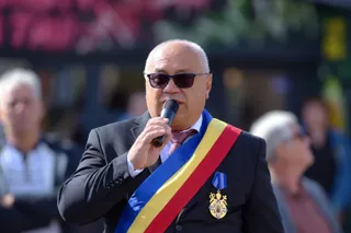 Un bărbat cu ochelari de soare și o bandă tricoloră pe piept vorbește într-un microfon în fața unor persoane desfocate.