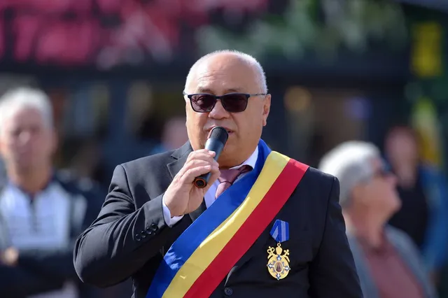 Un bărbat cu ochelari de soare și o bandă tricoloră pe piept vorbește într-un microfon în fața unor persoane desfocate.