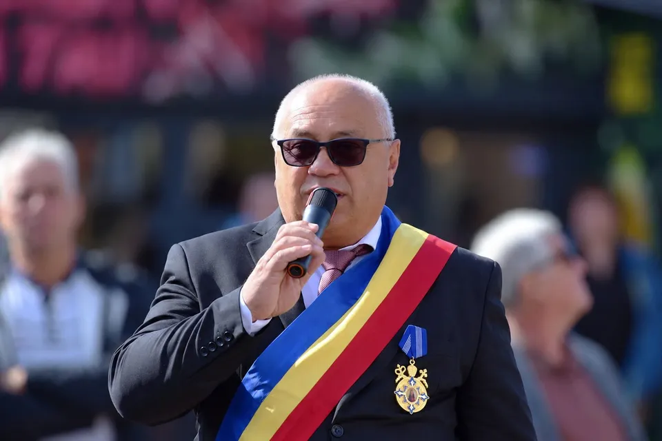 Un bărbat cu ochelari de soare și o bandă tricoloră pe piept vorbește într-un microfon în fața unor persoane desfocate.