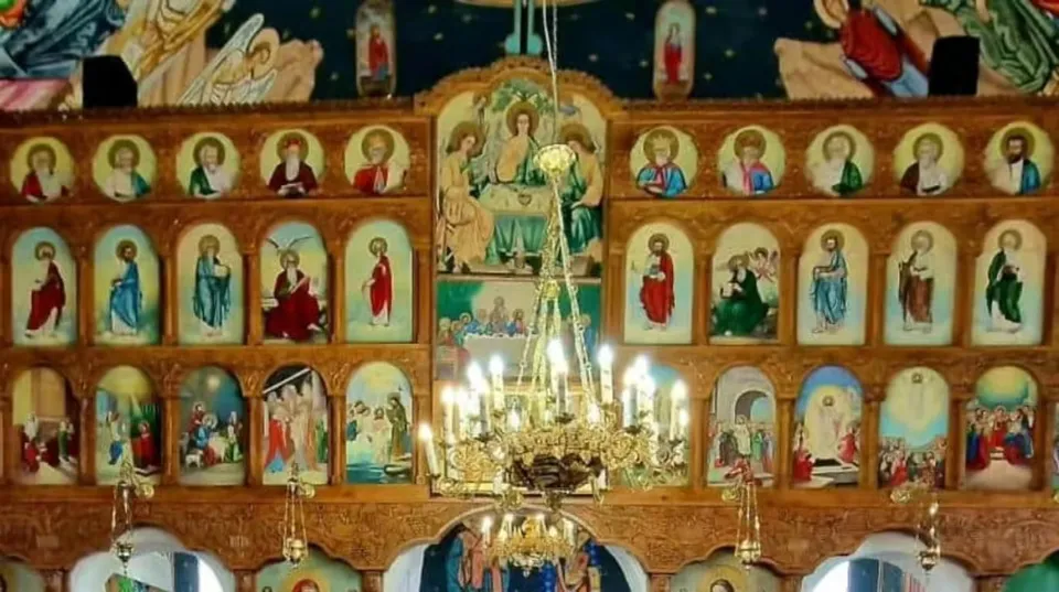 Un iconostas din lemn cu numeroase picturi religioase de tip icoană dispuse în mai multe rânduri, sub un candelabru metalic suspendat.