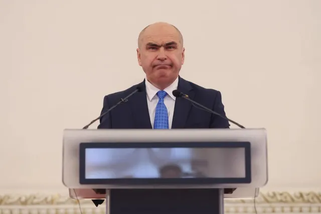Un bărbat în costum bleumarin și cravată albastră, cu părul încălzit pe cap, vorbește în picioare din spatele unui pupitru gri cu două microfoane.