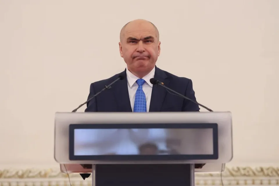 Un bărbat cu părul puțin, purtând sacou închis la culoare, cămașă albă și cravată albastră, stă în spatele unui podium cu două microfoane.