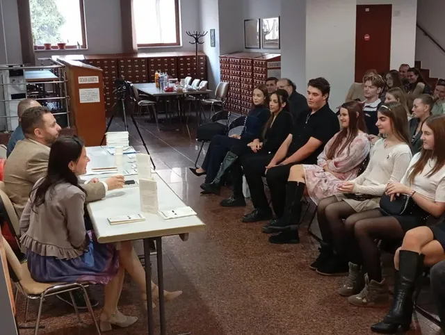 Un grup de persoane stă așezat pe scaune în fața unei mese albe pe care se află cărți și documente, într-o încăpere cu rafturi de depozitare.