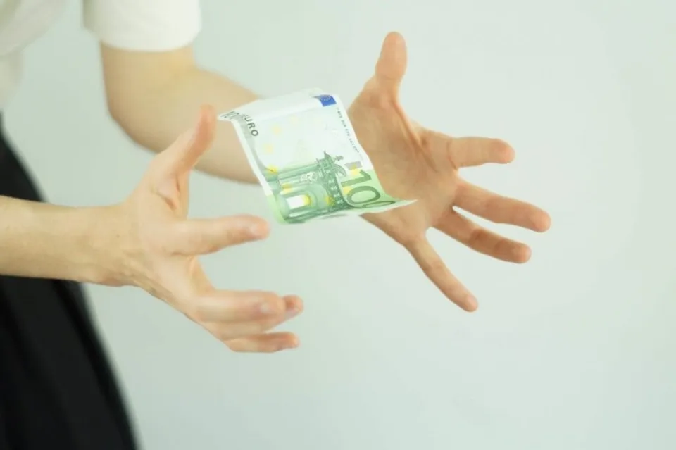 Două mâini întinse spre o bancnotă de zece euro care plutește în aer pe un fundal alb.