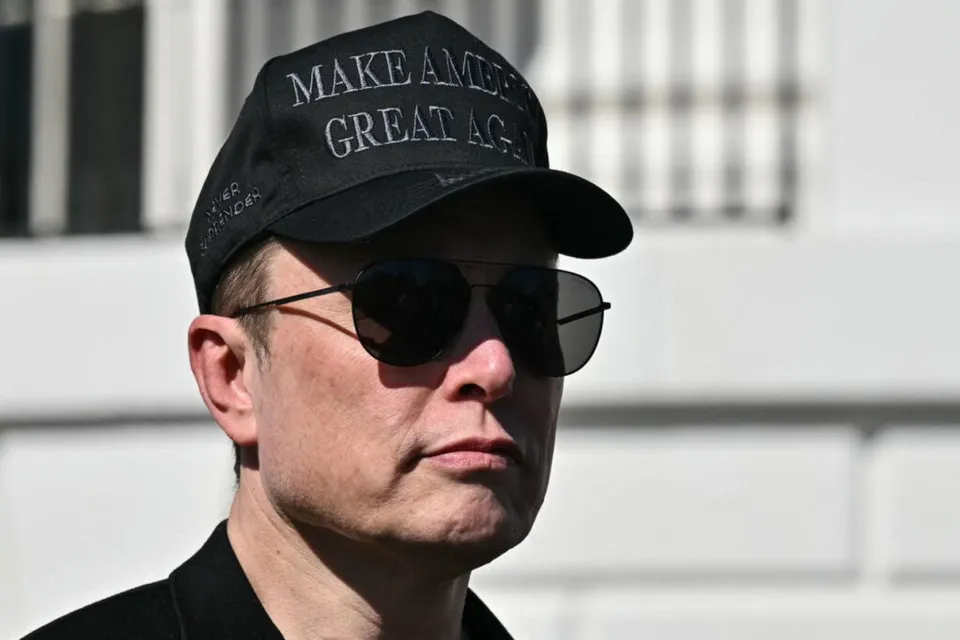 A man wearing a black cap with white text, dark sunglasses, and a black shirt is looking slightly to the side against a blurred white building background.