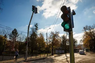Un semafor cu lumină verde este montat pe un stâlp metalic în prim-plan, sub un cer cu nori, lângă o stradă cu mașini și copaci.